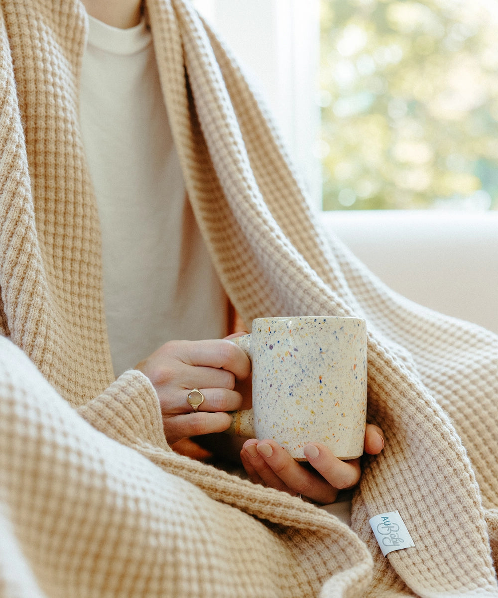 A close up of a woman sitting with a mug of tea, wrapped in a textured AU Baby throw blanket in Henna.