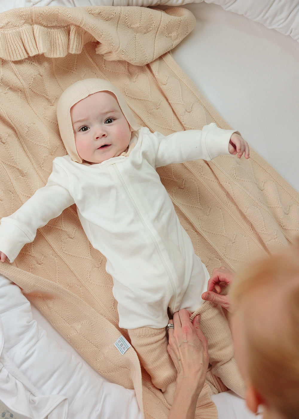 Baby in a white outfit lying on an AU Baby Butter Cream beige baby blanket with a person's hand visible.