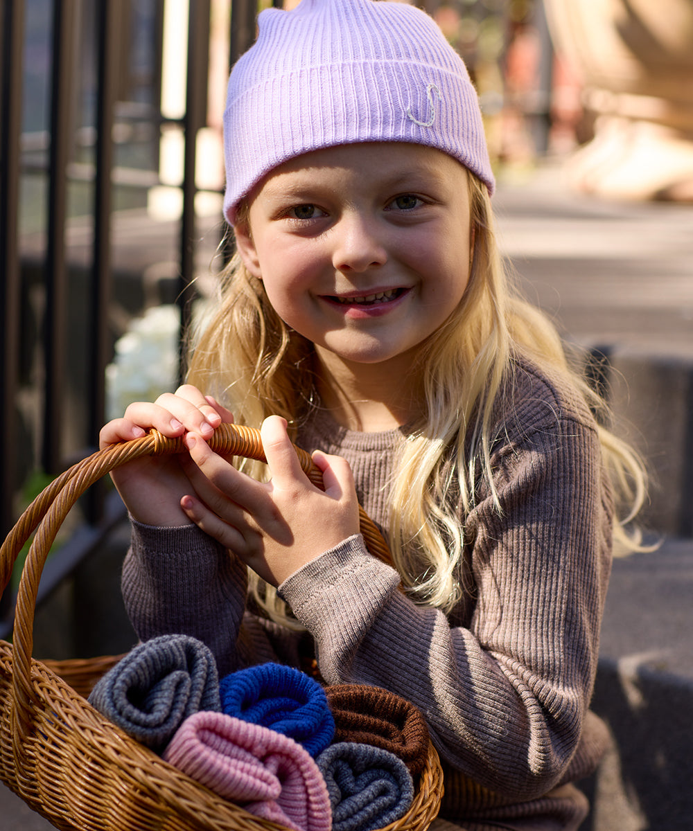 Close-up of a smiling girl wearing an AU Baby Lavender merino wool and cashmere beanie personalized with an embroidered initial, holding a basket with other beanies.